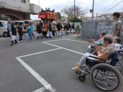 車椅子利用者を含む入居者が地域の祭りを観覧している風景